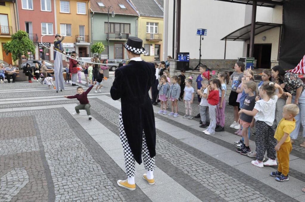 Szczudlarz i mężczyzna w cyrkowym stroju obracają linę. W środku chłopiec w pozie przygotowanej do skoku. Obok zgromadzone dzieci. W tle fasady okolicznych budynków.
