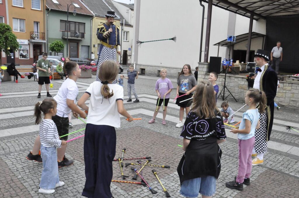 Szczudlarz, mężczyzna w cyrkowym stroju i grupka dzieci stoją w okręgu i podrzucają cyrkowymi pałeczkami na wybrukowanym placu. W tle fasady okolicznych budynków.