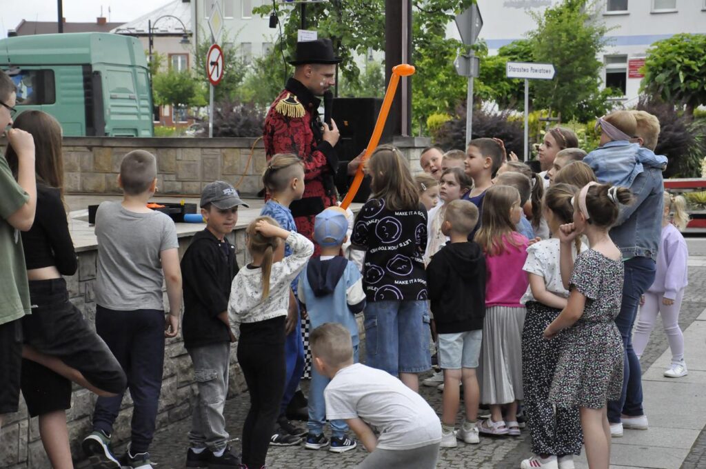 Duża grupa dzieci zgromadzona wokół cyrkowca, który trzyma długi balon. Wszyscy stoją obok sceny plenerowej. W tle zieleń miejskiego parku.