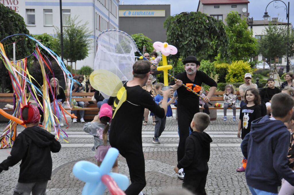 Dwoje mimów w czarnych strojach trzyma rekwizyty artystyczne. Wokół nich biegają uśmiechnięte dzieci. W tle zieleń miejskiego parku.