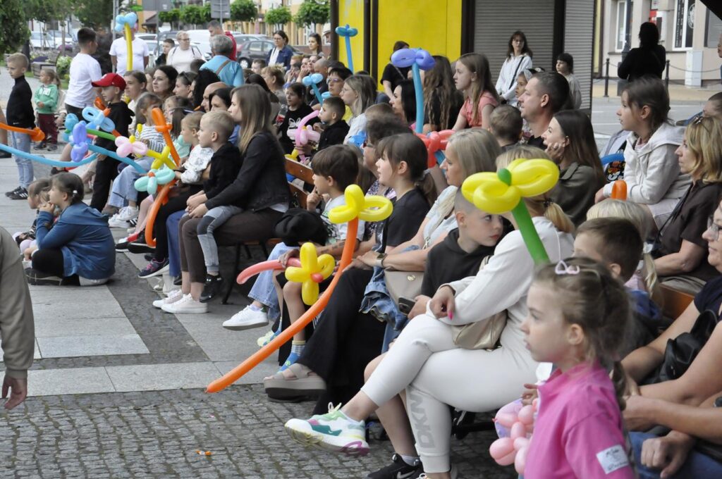 Duża grupa dzieci i dorosłych. Niemal wszyscy siedzą. Niektórzy trzymają kolorowe balony w formie kwiatów. W tle miejska ulica.