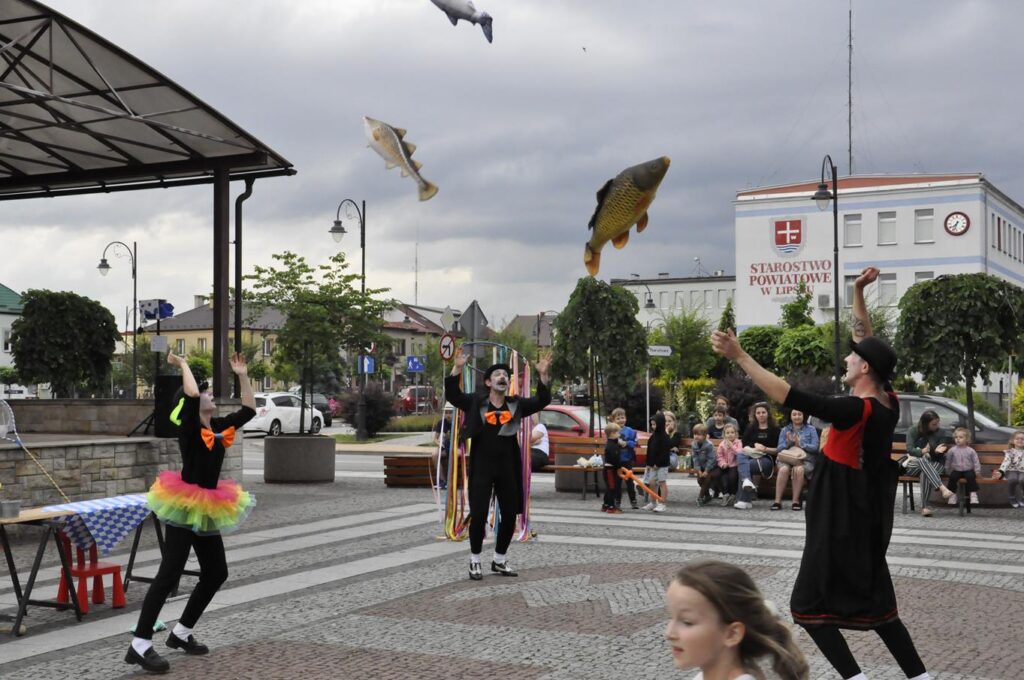 Troje mimów w czarnych strojach podrzuca maskotki w kształcie ryb. W tle kilkoro dorosłych i dzieci, którzy siedzą na ławkach. Dalej zieleń miejskiego parku oraz fasady okolicznych budynków.