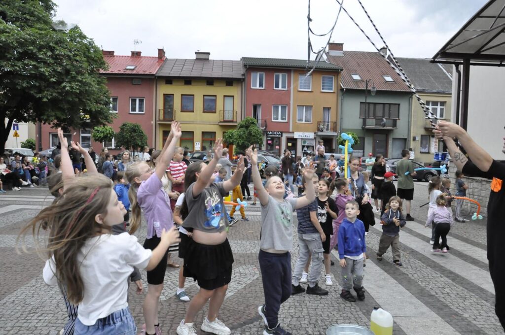 Duża grupa uśmiechniętych dzieci. Niektórzy unoszą ręce do góry. W tle kolorowe fasady okolicznych budynków.