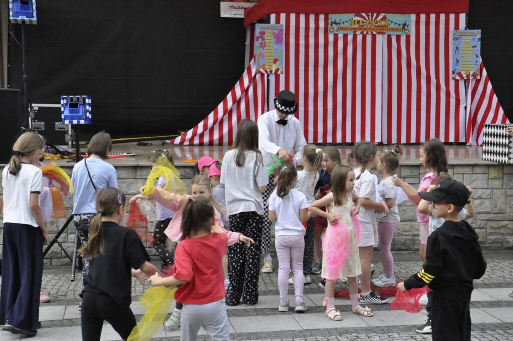 Dzieci w kolorowych zwykłych ubraniach zgromadzone wokół mężczyzny przebranego za cyrkowca. Za nimi scena plenerowa na której stoi namiot cyrkowy.