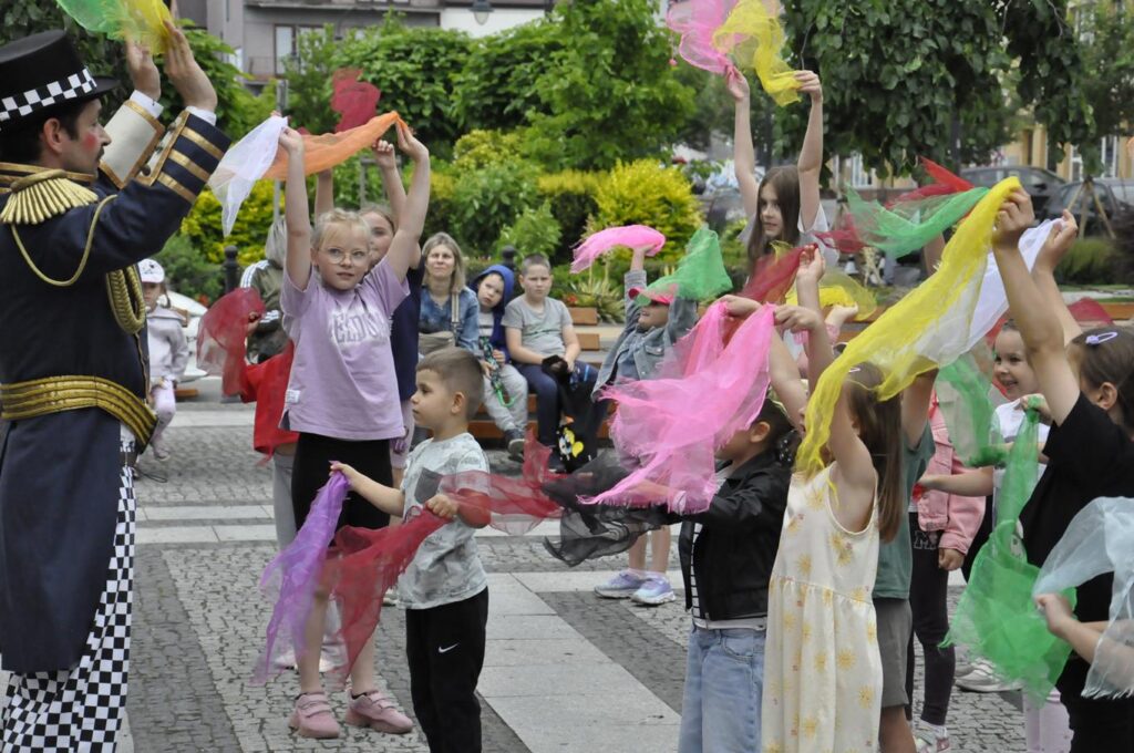 Dzieci wymachują kolorowymi chustkami. W tle zieleń miejskiego parku.