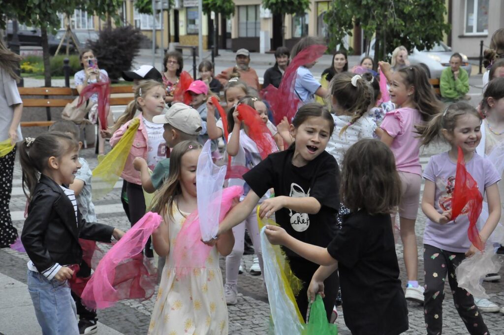 Dzieci wymachują kolorowymi chustkami. W tle miejska ulica.