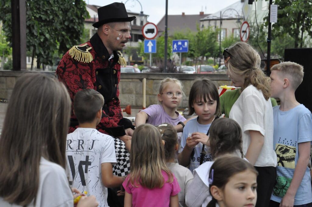 Dzieci zgromadzone wokół cyrkowca w czarnym kapeluszu. W tle zieleń miejskiego parku.