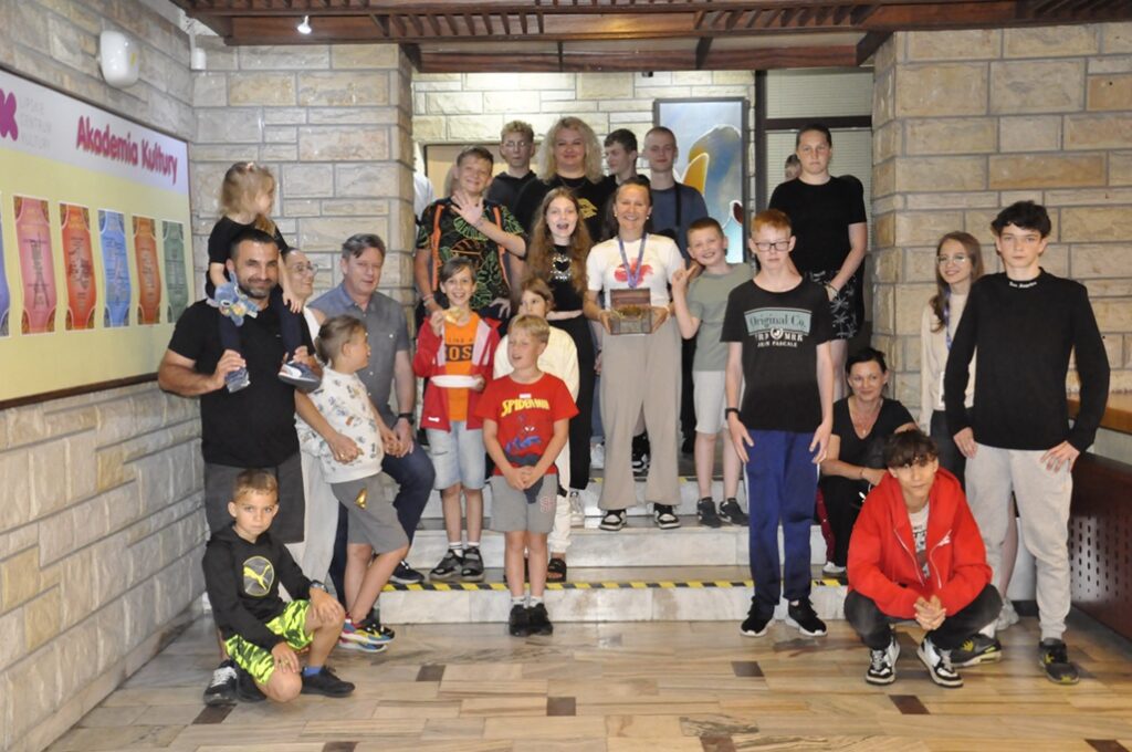 Duża grupa młodzieży i dorosłych stoi na schodkach w holu budynku. Dziewczynka w centrum trzyma otwartą szkatułkę. Pomieszczenie ciepło oświetlone.