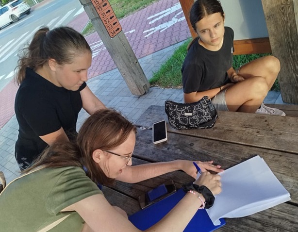 Trzy dziewczynki stoją lub siedzą przy stole. Jedna z nich zapisuje coś na kartce. Plener, altana miejska.
