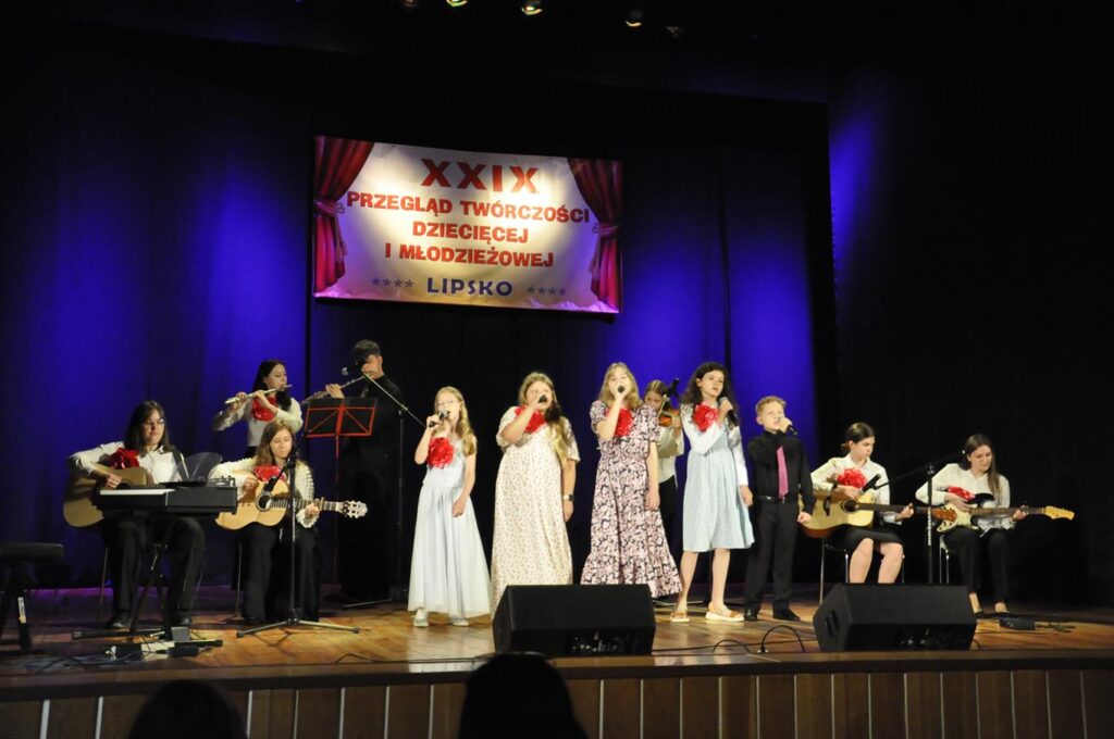 Grupa kilkunastu młodych osób instrumentaliści i wokaliści stoją lub siedzą na scenie. Stroje oficjalne. Nad nimi baner z nazwą wydarzenia.