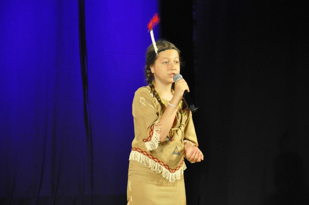 Dziewczynka w stroju indianki trzyma mikrofon przy ustach. Tło czarno-granatowe.