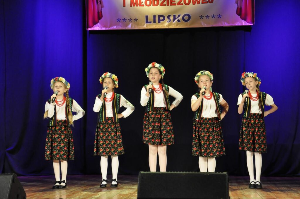 Pięć dziewczynek w ludowych strojach i wiankami na głowach trzyma mikrofony. Tło czarno-granatowe. Nad nimi fragment baneru z nazwą wydarzenia.
