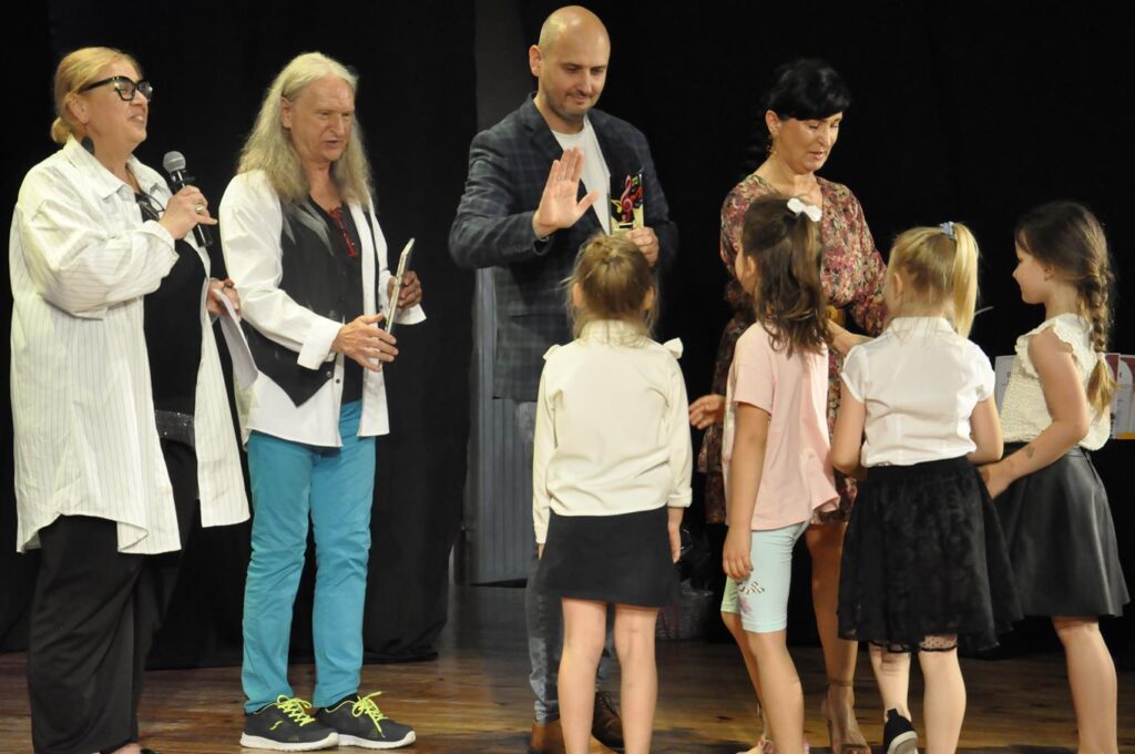 Czworo dorosłych, kobiety i mężczyźni przybijają "piątkę" dzieciom. Kobieta w białej koszuli trzyma mikrofon. Tło czarne.