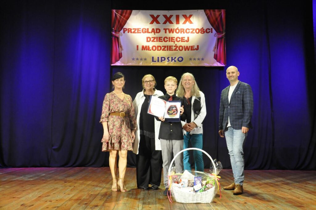 Chłopiec w czarnej koszuli trzyma etui ze statuetką Grand Prix. Za nim stoi kilku dorosłych, przed nim znajduje się kosz ze słodyczami. Nad nimi baner z nazwą wydarzenia.