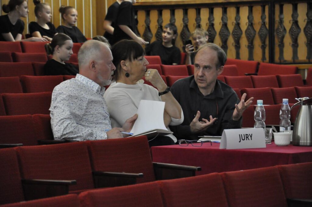 Troje dorosłych: kobieta i dwóch mężczyzn obraduje przy stole z kartką na której widnieje napis "JURY". Wokół czerwone fotele, niektóre zajęte przez młodzież w czarnych strojach. Pomieszczenie ciepło oświetlone.