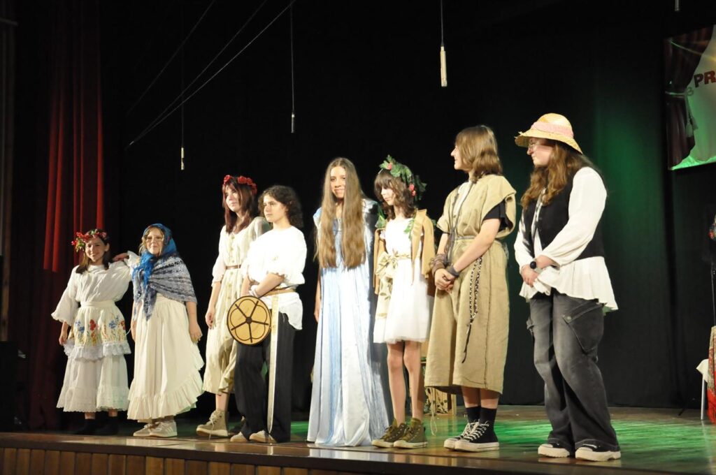 Grupa młodzieży w scenicznych strojach w klimacie folkloru stoi na scenie. Tło czarne.