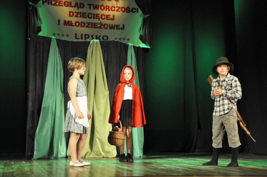 Troje dzieci stoi na scenie. Postać myśliwego, czerwonego kapturka i osoba w białym fartuszku. Za nimi element scenografii przypominający las. W tle baner z nazwą wydarzenia.