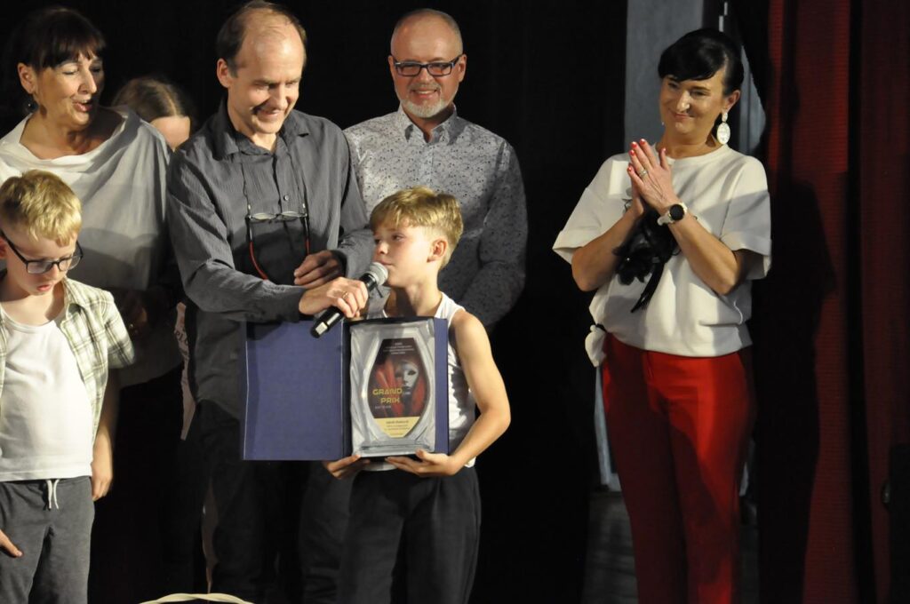 Kilka osób. Chłopiec trzyma etui ze statuetką GRAND PRIX. Mężczyzna w szarej koszuli przykłada mu do ust mikrofon. Tło szaro-czarne.
