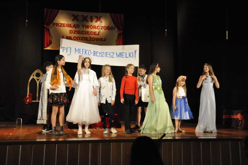 Dzieci w kolorowych strojach scenicznych, szlacheckich i baśniowych. Wszyscy stoją stoją. Niektóre dzieci trzymają transparent z napisem " Pij mleko - będziesz wielki!. Nad nimi baner z nazwą wydarzenia.