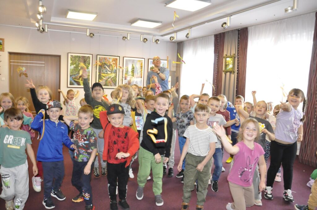 Duża grupa w kolorowych zwykłych ubraniach podskakuje i podrzuca żółte wstążki. Za nimi znajduje się uśmiechnięty mężczyzna w szarej koszuli i w okularach. Pomieszczenie ciepło oświetlone.
