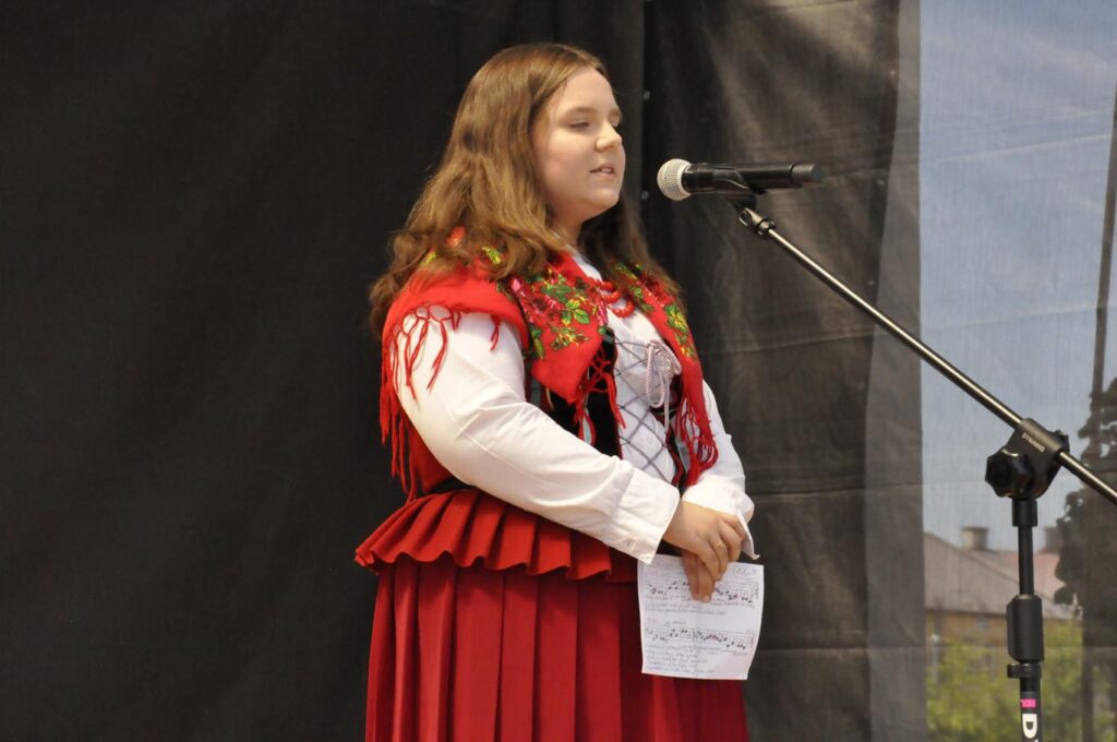 Dziewczyna w rozpuszczonych, brązowych i półdługich włosach oraz w czerwonym wzorzystym stroju stoi przy mikrofonie na statywie. Tło czarne. Pogodny dzień.