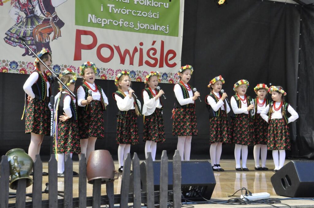 Grupa dziewczynek z wiankami na głowach i ludowych strojach stoją na scenie. Niektóre trzymają mikrofony przy ustach. Za nimi wisi baner z nazwą wydarzenia. Pogodny dzień.