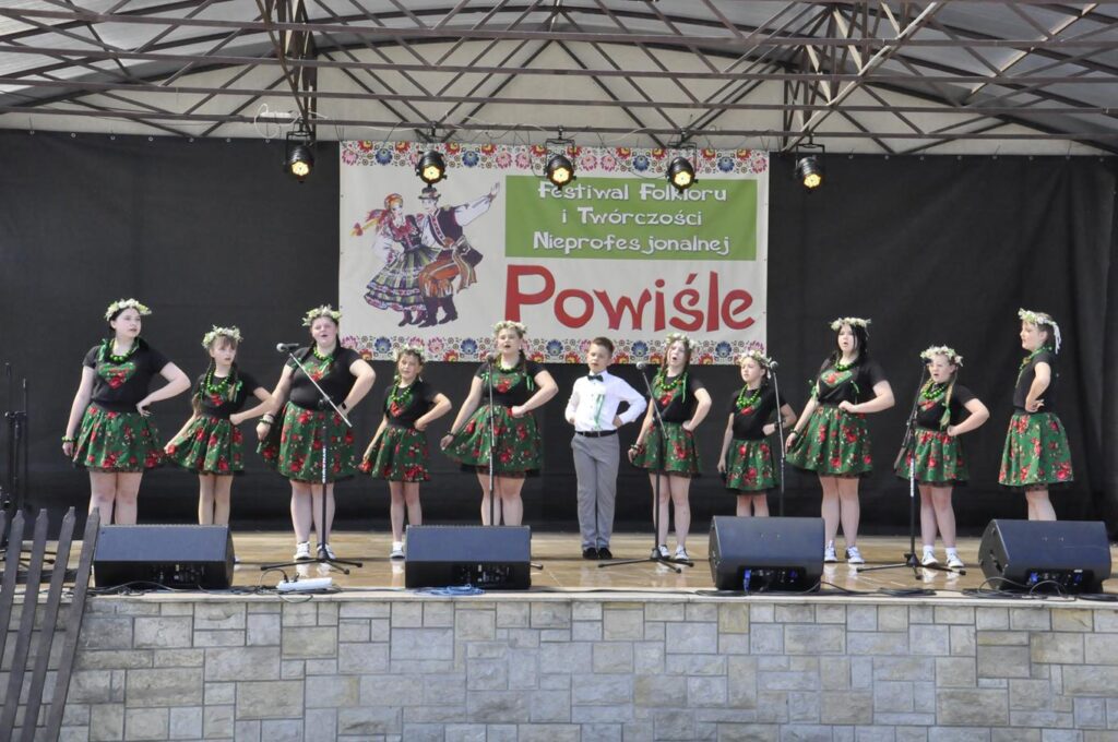 Grupa nastolatków w ludowych strojach stoją na scenie plenerowej. Dziewczynki mają wianki na głowach. Przed nimi stoją mikrofony na statywach. Za nimi baner z nazwą wydarzenia. Pogodny dzień.