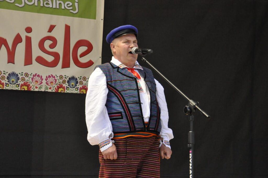 Mężczyzna w granatowo-czerwonym ludowym stroju stoi przy mikrofonie na statywie. Za nim fragment baneru promującego wydarzenie. Pogodny dzień.