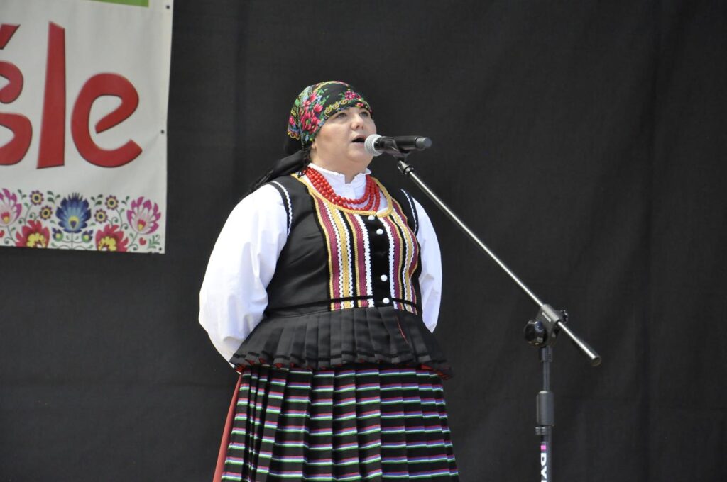 Kobieta w kolorowej chuście na głowie i ludowym stroju stoi przy mikrofonie na statywie. Za nią fragment baneru promującego wydarzenie. Pogodny dzień.