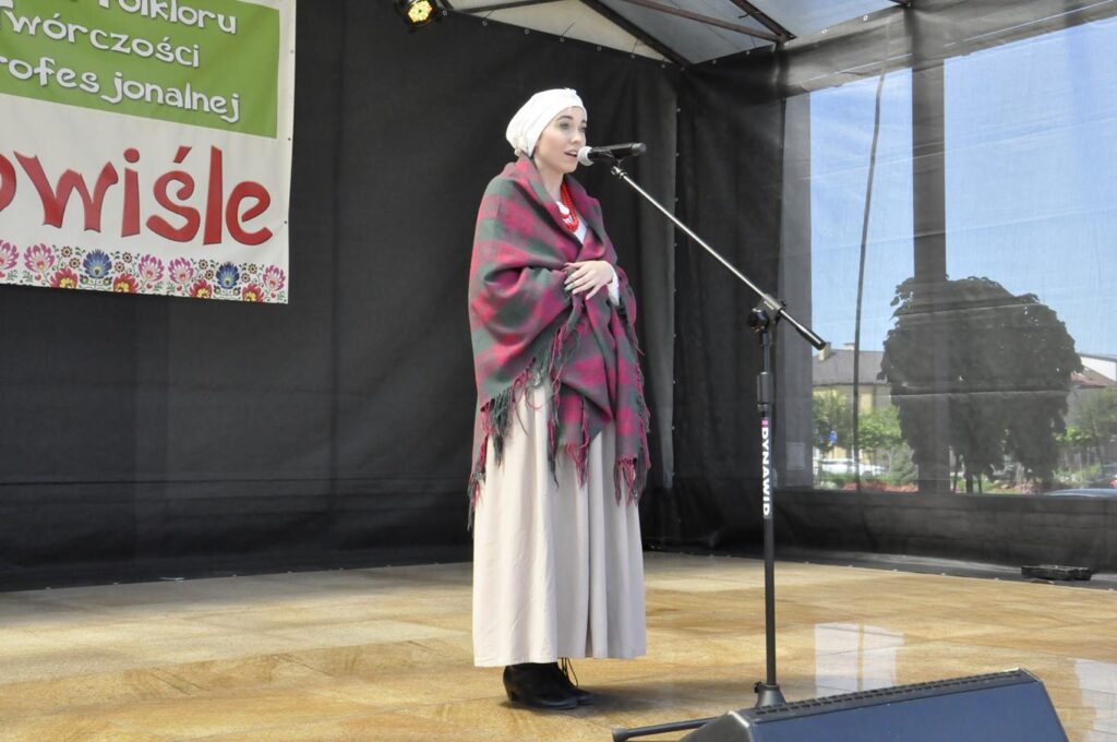 Młoda kobieta w białej chuście na głowie, w szarej spódnicy i kraciastej długiej chuście okrywającej ramiona stoi na scenie plenerowej przy mikrofonie na statywie. Za nią fragment baneru promującego wydarzenie. Pogodny dzień.