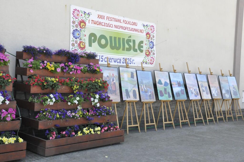 Piętrowa rabata z kolorowymi kwiatami. Obok wystawa galerii regionalnej zdjęć strojów ludowych w antyramach na sztalugach stojących w rzędzie. Nad galerią baner wiszący na elewacji budynku i promujący wydarzenie.