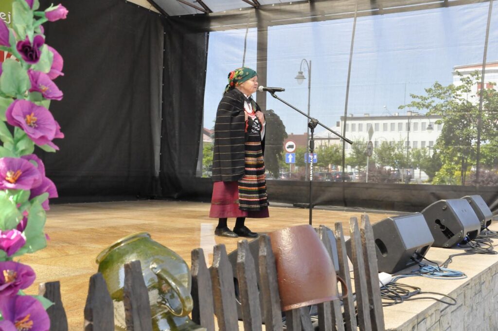 Kobieta w zielonej chuście z motywem kwiatów i w ludowym stroju stoi na scenie plenerowej przy mikrofonie na statywie. Pogodny dzień.