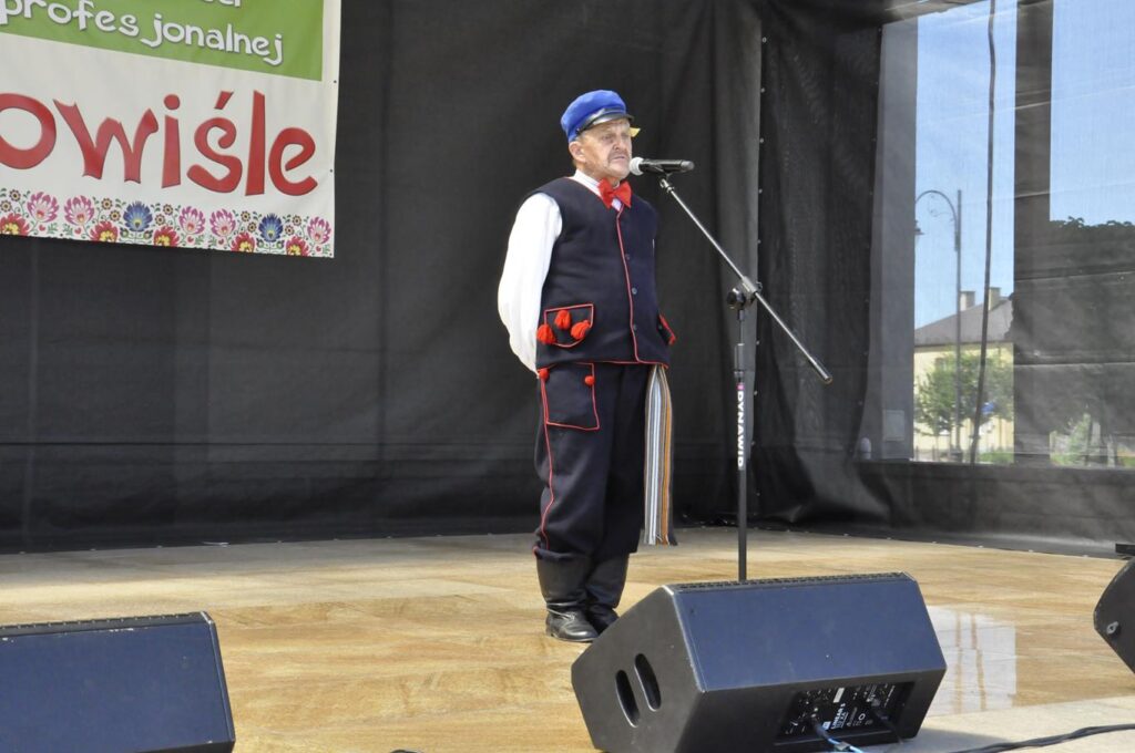 Mężczyzna w niebieskim kaszkiecie i ludowym stroju stoi na scenie plenerowej przy mikrofonie na statywie. Za nim fragment baneru promującego wydarzenie. Pogodny dzień.