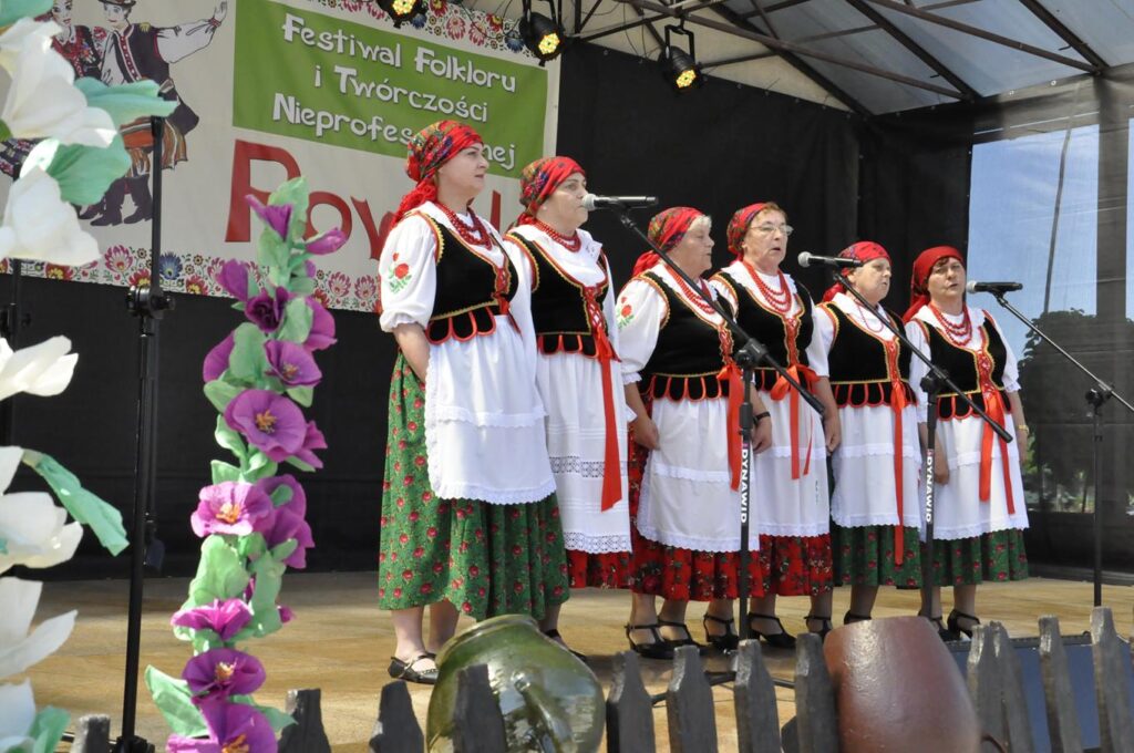 Sześć kobiet w czerwonych chustach na głowie i ludowych strojach z białymi fartuchami stoi na scenie plenerowej przy kilku mikrofonach na statywach. Za nimi wisi baner promujący wydarzenie.