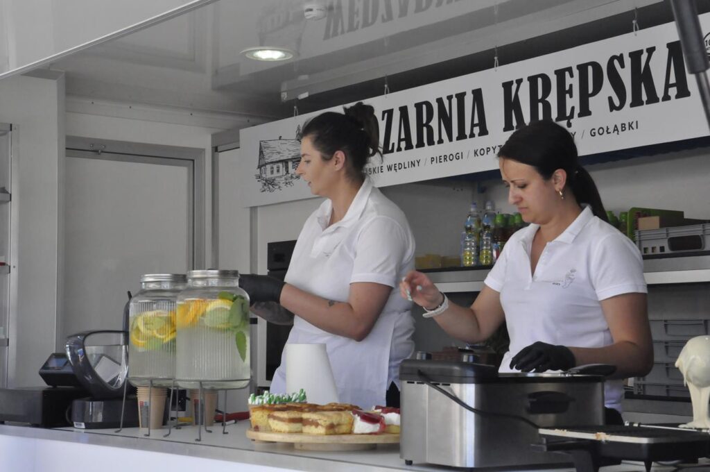 Dwie kobiety w białych koszulkach z kołnierzykami we wnętrzu Foodtracka. Nad nimi wisi baner z nazwą stoiska.
