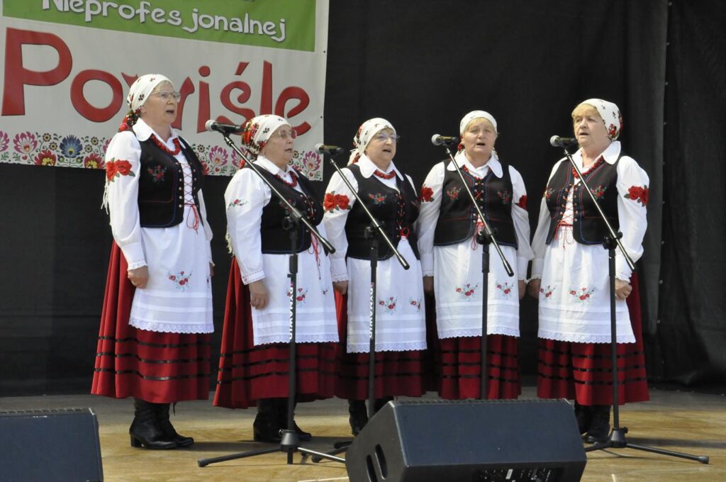 Pięć kobiet w białych chustach na głowie i ludowych strojach stoi na scenie plenerowej przy kilku mikrofonach na statywach. Za nimi fragment baneru promującego wydarzenie. Pogodny dzień.