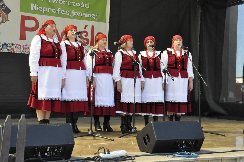 Sześć kobiet w czerwonych chustach na głowie i czerwono-białych ludowych strojach stoi na scenie plenerowej przy kilku mikrofonach na statywach. Za nimi fragment baneru promującego wydarzenie. Pogodny dzień.