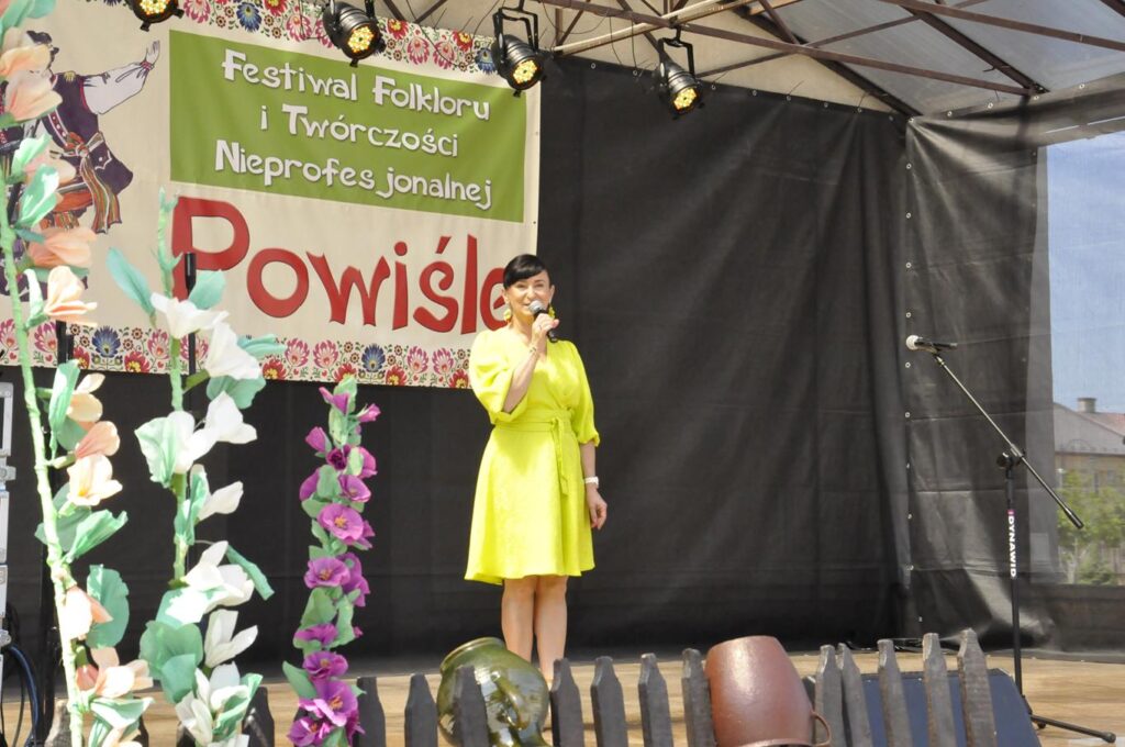 Kobieta w żółtej sukience stoi na scenie plenerowej i trzyma mikrofon przy ustach za nie baner promujący wydarzenie. Pogodny dzień.
