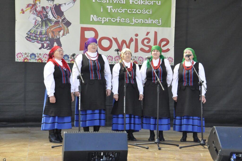 Pięć kobiet w różnokolorowych chustach na głowie i ludowych strojach stoi na scenie plenerowej przy kilku mikrofonach na statywach. Za nimi wisi baner promujący wydarzenie. Pogodny dzień.