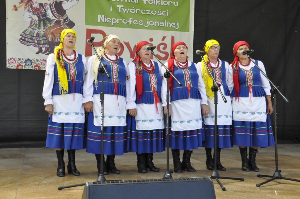 Sześć kobiet w kolorowych chustach na głowie i ludowych niebiesko-białych strojach stoi na scenie plenerowej przy kilku mikrofonach na statywach. Za nimi fragment baneru promującego wydarzenie. Pogodny dzień.