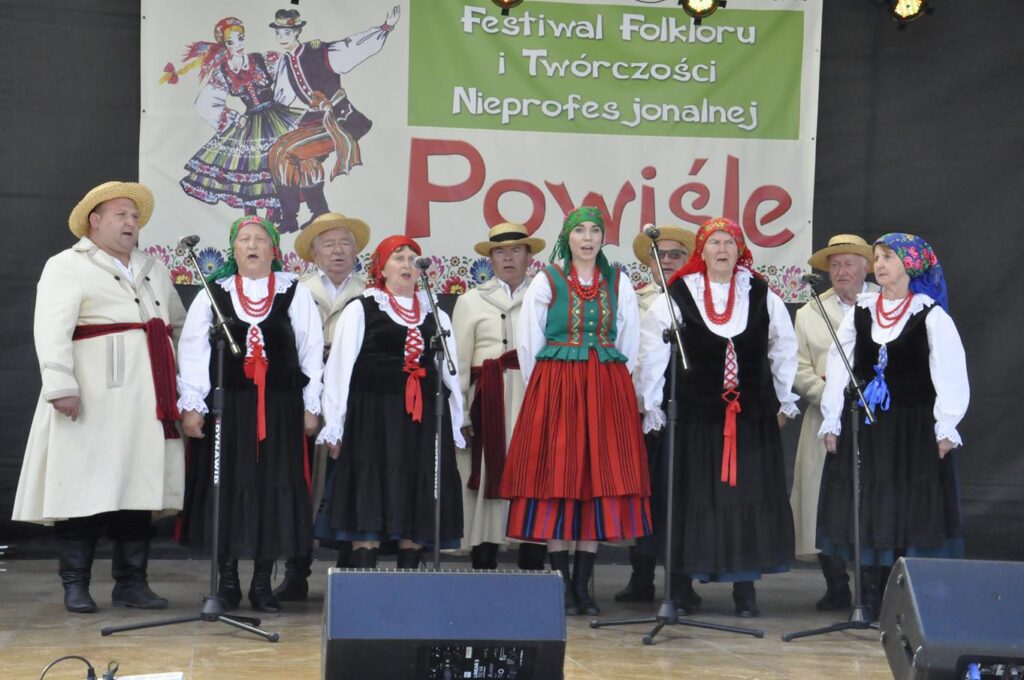 Grupa śpiewaków (kobiety i mężczyźni ludowych strojach stoi na scenie plenerowej przy kilku mikrofonach na statywach. Mężczyźni mają na głowach słomiane kapelusze a kobiety kolorowe chusty.Za nimi fragment baneru promującego wydarzenie. Pogodny dzień.