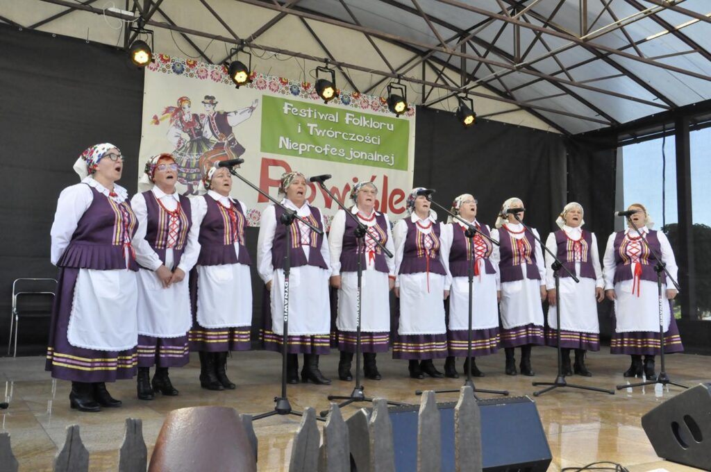 Grupa kilkunastu kobiet w jasnych, kwiatowych chustach na głowie i ludowych fioletowo-białych strojach stoi na scenie plenerowej przy kilku mikrofonach na statywach. Za nimi fragment baneru promującego wydarzenie. Pogodny dzień.