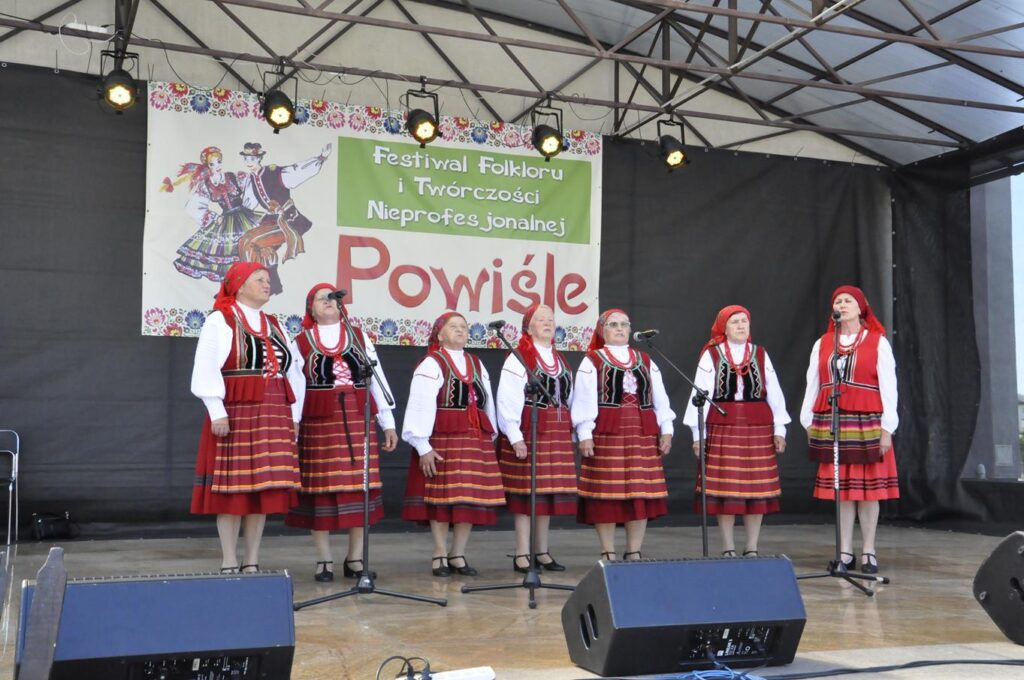 Siedem kobiet w czerwonych chustach na głowie i ludowych czerwonych strojach w pasy stoi na scenie plenerowej przy kilku mikrofonach na statywach. Za nimi fragment baneru promującego wydarzenie. Pogodny dzień.