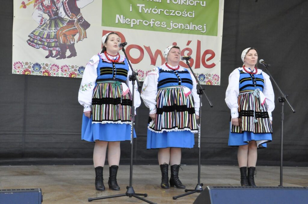 Trzy kobiety w białych chustach na głowie i ludowych błękitno-białych strojach z pasiastymi fartuchami stoi na scenie plenerowej przy kilku mikrofonach na statywach. Za nimi fragment baneru promującego wydarzenie. Pogodny dzień.