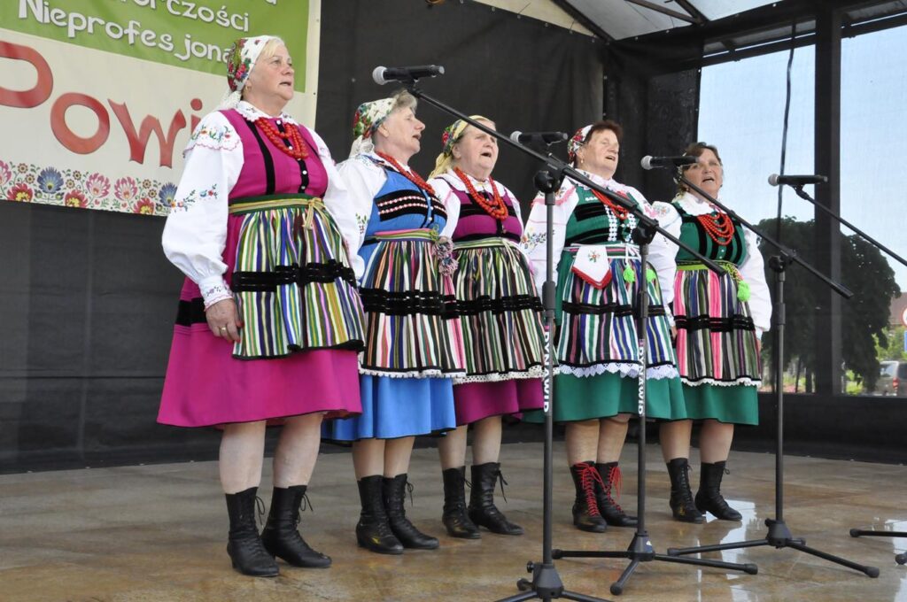Pięć kobiet w kwiatowych chustach i w kolorowych ludowych strojach stoi na scenie plenerowej przy kilku mikrofonach na statywach. Za nimi fragment baneru promującego wydarzenie. Pogodny dzień.