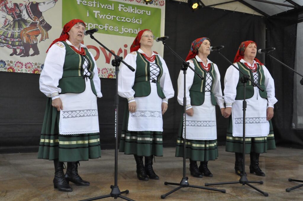 Cztery kobiety w czerwonych chustach na głowie i ludowych zielono-białych strojach stoi na scenie plenerowej przy kilku mikrofonach na statywach. Za nimi fragment baneru promującego wydarzenie. Pogodny dzień.