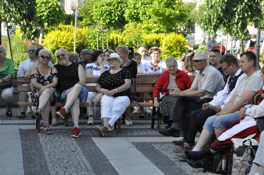 Grupa ludzi w zwykłych, kolorowych ubraniach siedzi na ławkach. w Tle parkowa zieleń. Pogodny dzień.