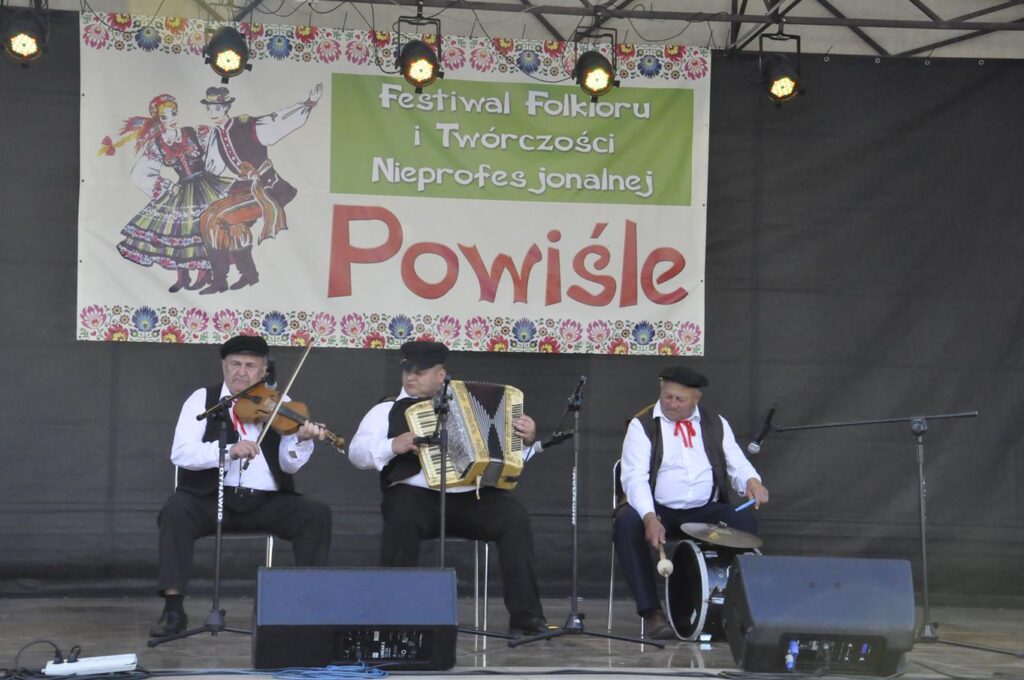 Trzech mężczyzn w ludowych strojach siedzi na krzesłach na scenie plenerowej. Jeden gra na skrzypcach, drugi na akordeonie, a trzeci na bębenku. Za nimi wisi baner promujący wydarzenie.