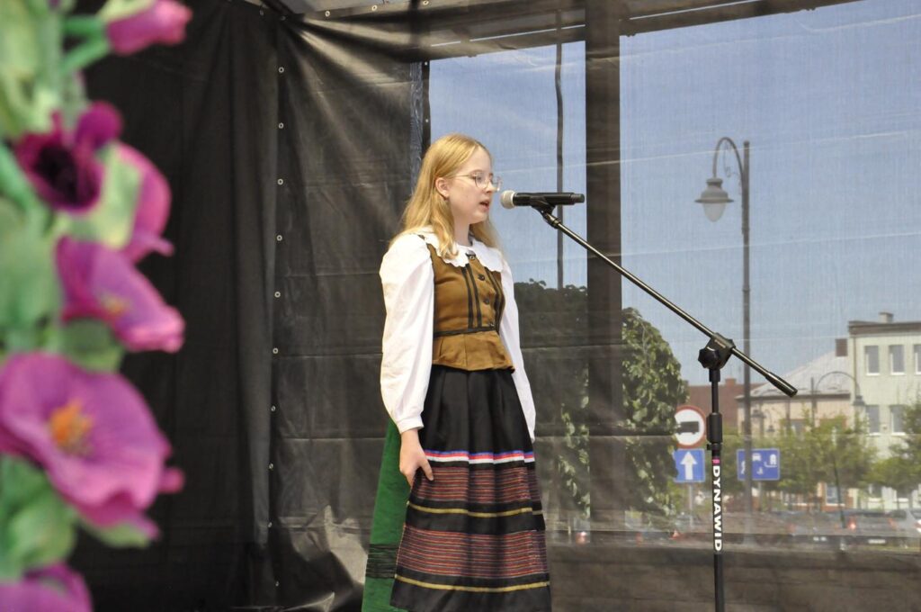 Dziewczyna w w blond półdługich włosach w okularach i stroju ludowym stoi przy mikrofonie na statywie. tło czarne. Pogodny dzień.