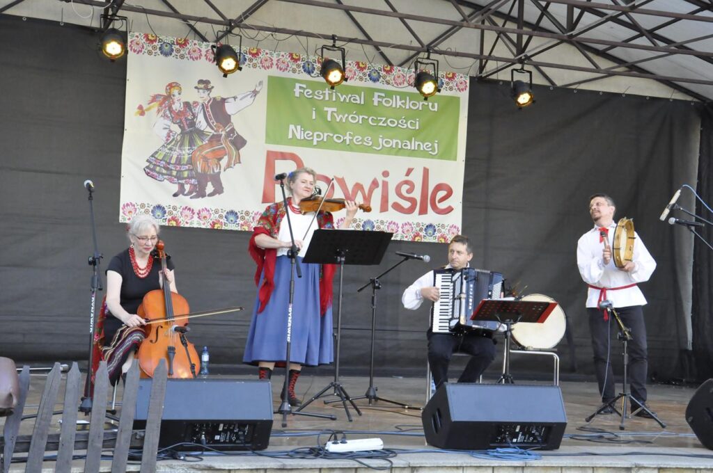 Grupa muzyków w ludowych strojach. Niektórzy stoją, inni siedzą. Jedna kobieta gra na wiolonczeli, druga na skrzypcach. Jeden mężczyzna gra na akordeonie, a drugi na tamburynie. Za nimi wisi baner promujący wydarzenie.
